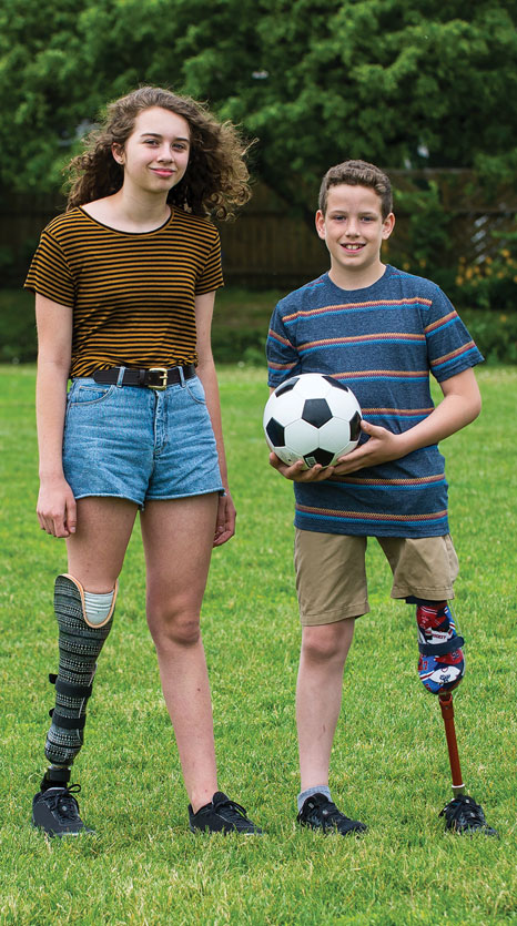 A teenage female leg amputee stands next to a younger male leg amputee on a soccer field.