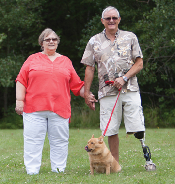A senior male leg amputee walks with his wife and small dog in a grassy field.