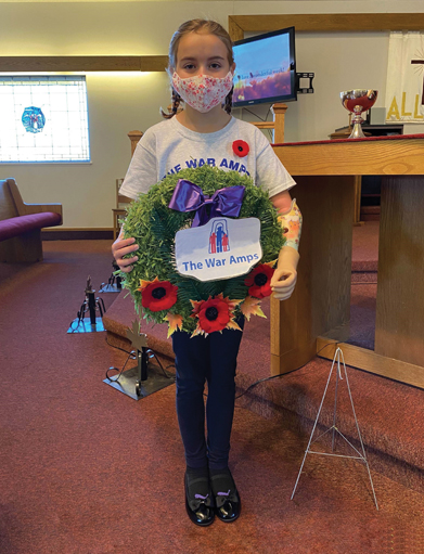 A young female arm amputee holds a Remembrance Day wreath with The War Amps written on it while wearing a cloth face mask.