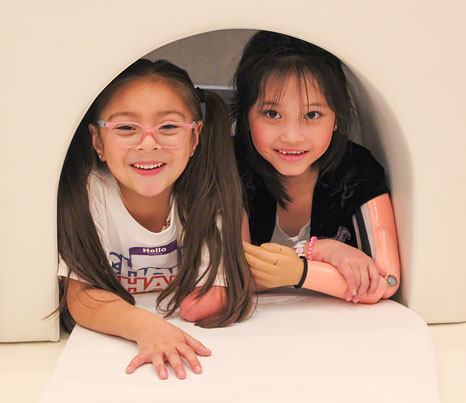 Two young girls, both arm amputees, play inside a white foam structure.