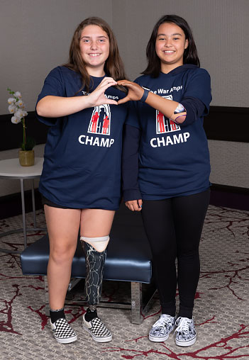Two young girls, one an arm amputee and the other a leg amputee, join their hands together to create the shape of a heart.